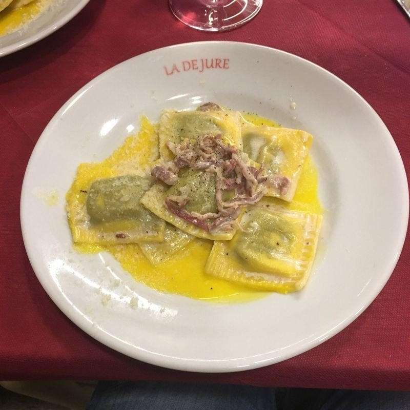 Ravioli di patate viola con burro fuso e speck croccante, nel piatto con la scritta La De Jure.
