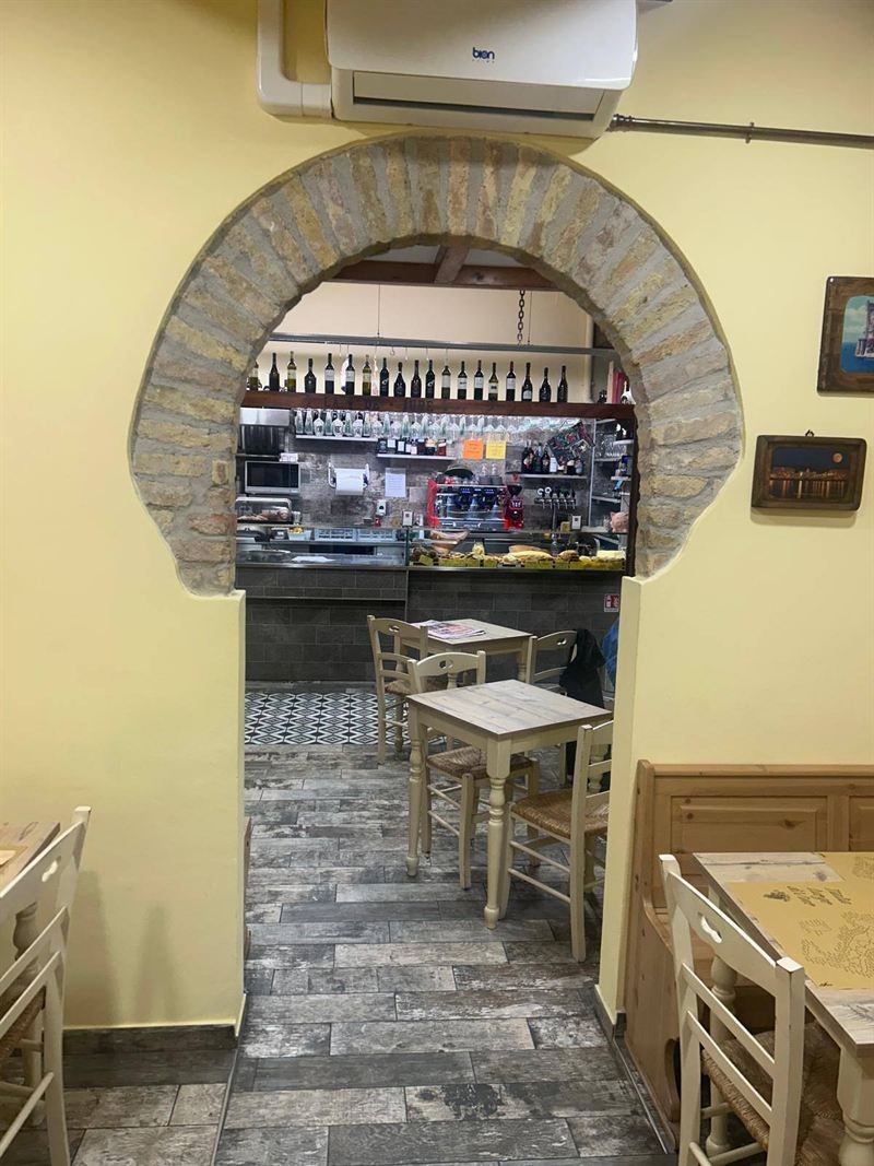 La sala con le pareti gialle, sedie impagliate e un arco in mattoni che si apre sul banco con le bottiglie.